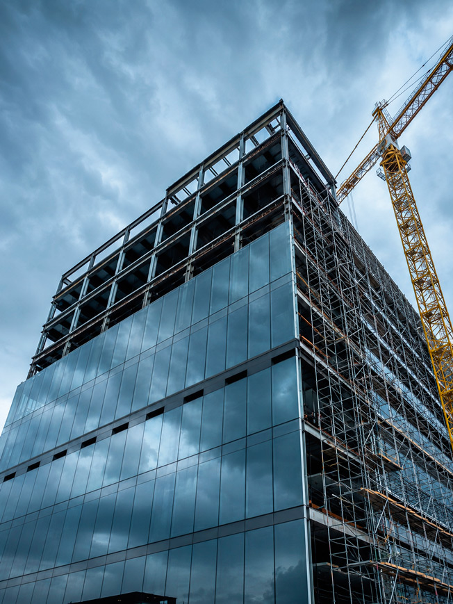 Fotografía arquitectónica real de edificio corporativo moderno en construcción avanzada en Ciudad de México: vista en plano contrapicado a 45° desde la banqueta mirando hacia arriba; fachada de cristal y aluminio anodizado gris oscuro reflejando el cielo azul acero nublado; estructura metálica de vigas IPR color gris acero expuesta en los últimos dos pisos superiores aún sin cerrar; andamiaje de aluminio plateado en el costado derecho; grúa torre de color amarillo industrial al fondo derecho; cielo azul acero dramático con nubes gris oscuro; paleta: azul acero del cielo, gris oscuro aluminio fachada, amarillo grúa, plateado andamiaje, negro de las sombras profundas entre vigas; composición dinámica en contrapicado extremo con gran angular; sin texto visible, sin logotipos; iluminación natural de cielo nublado que genera luz difusa uniforme; estilo fotografía arquitectónica editorial industrial de alta producción.