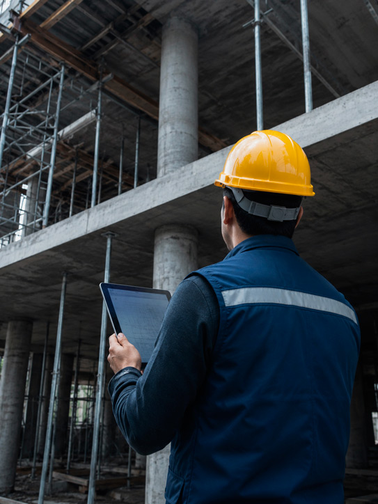 Fotografía documental real de supervisión técnica de obra en Ciudad de México: ingeniero civil con casco amarillo brillante y chaleco azul marino de espaldas, parado frente a estructura de concreto armado con columnas de gran diámetro y losa en proceso de construcción; sostiene tableta digital con planos digitales; fondo de estructura de concreto gris con encofrado de acero plateado; luz natural difusa lateral que ilumina el casco amarillo como único punto de color vivo; entorno cromático de grises azulados — gris oscuro concreto, gris plateado acero, azul marino chaleco — con el amarillo del casco como único acento; composición de plano medio trasero sin rostro identificable; sin texto legible, sin logotipos; estilo fotografía documental editorial de obra civil industrial.