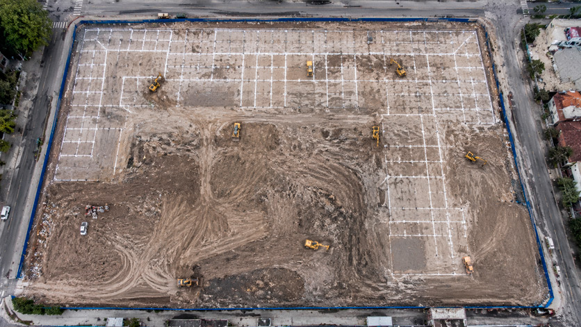 Fotografía aérea desde drone de preparación de terreno urbano en CDMX: vista cenital a 70m de altura de predio de gran extensión con suelo arcilloso café gris en distintas etapas de nivelación; trazos de cimentación en cal blanca formando retícula ortogonal visible desde arriba como dibujo geométrico abstracto; perímetro del predio delimitado con malla de obra azul marino; maquinaria de terracería de color amarillo industrial distribuida en distintos puntos del predio; calles perimetrales asfaltadas gris oscuro; paleta: gris-café suelo arcilloso, blanco trazos de cal, azul marino malla perimetral, amarillo maquinaria, gris asfalto; composición cenital geométrica abstracta; sin texto, sin logotipos; estilo fotografía aérea documental de obra civil de alta producción.
