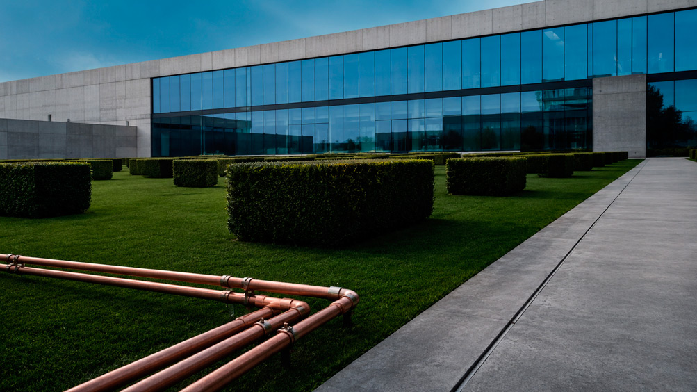 Fotografía de jardín corporativo de diseño contemporáneo y estética industrial: espacio verde de exterior de conjunto de oficinas con césped verde oscuro intenso perfectamente nivelado; arbustos ornamentales de podocarpus recortados en prismas rectangulares perfectos distribuidos en cuadrícula regular; tuberías de cobre del sistema de riego tecnificado visibles en primer plano como elemento gráfico industrial; pavimento de concreto industrializado gris con junta de expansión como sendero; fachada de edificio industrial moderno de concreto expuesto y cristal azul como telón de fondo; cielo azul acero; paleta: verde oscuro intenso del césped, cobre de las tuberías de riego, gris concreto pavimento, azul acero cristal edificio; sin personas; estilo fotografía de paisajismo industrial moderno de alta calidad.